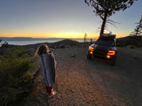 A person and dog watch a serene lake sunset, an adventure truck parked nearby. This view awaits your next RV rental adventure!