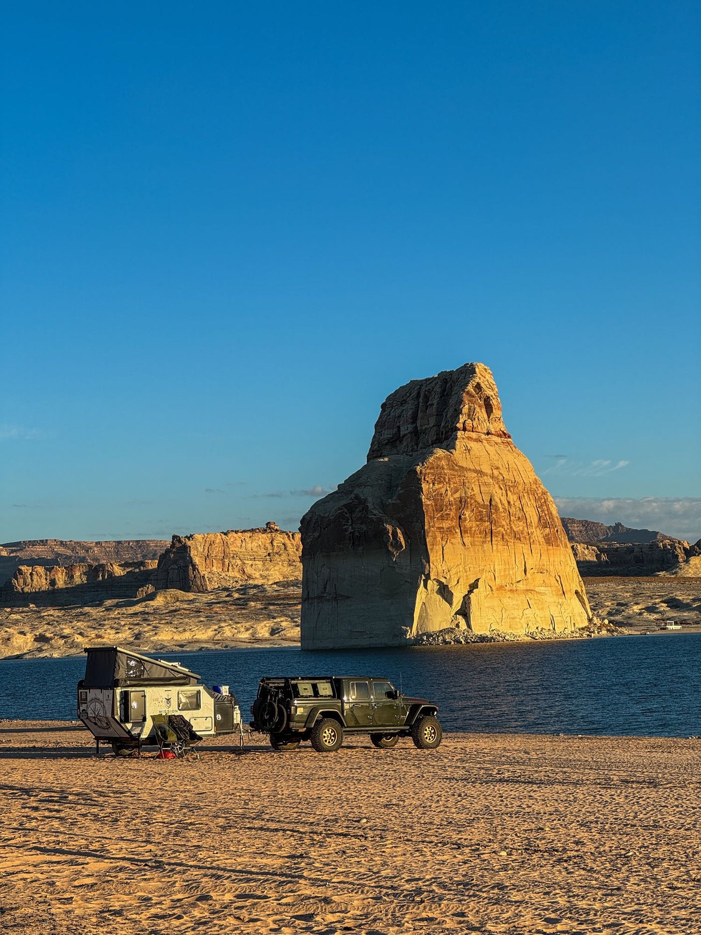 Scenic lakefront camping with an off-road RV rental and Jeep. A huge sunlit rock formation towers over the sandy beach campsite.