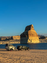 Scenic lakefront camping with an off-road RV rental and Jeep. A huge sunlit rock formation towers over the sandy beach campsite.