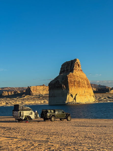 Scenic lakefront camping with an off-road RV rental and Jeep. A huge sunlit rock formation towers over the sandy beach campsite.