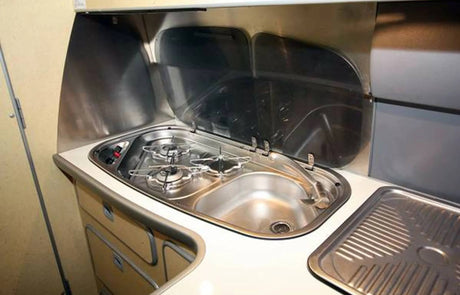 Compact RV rental kitchen featuring a two-burner stove, sink, and drainboard, perfect for cooking on your adventure.