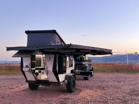 An overland camper RV rental is set up for off-grid adventure at dusk, featuring a pop-top tent and open mobile kitchen.