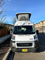 Front view of a white Winnebago camper van, an RV rental with its pop-top extended, parked on a sunny residential street in Oregon.