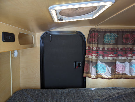 View inside a compact RV rental: wood-paneled interior, black door, ceiling fan/light, bear/fish curtain, and textured bed.