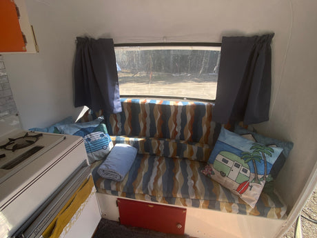 Cozy RV rental interior with a striped bench bed, camper pillows, and a compact stove. Views of nature outside.