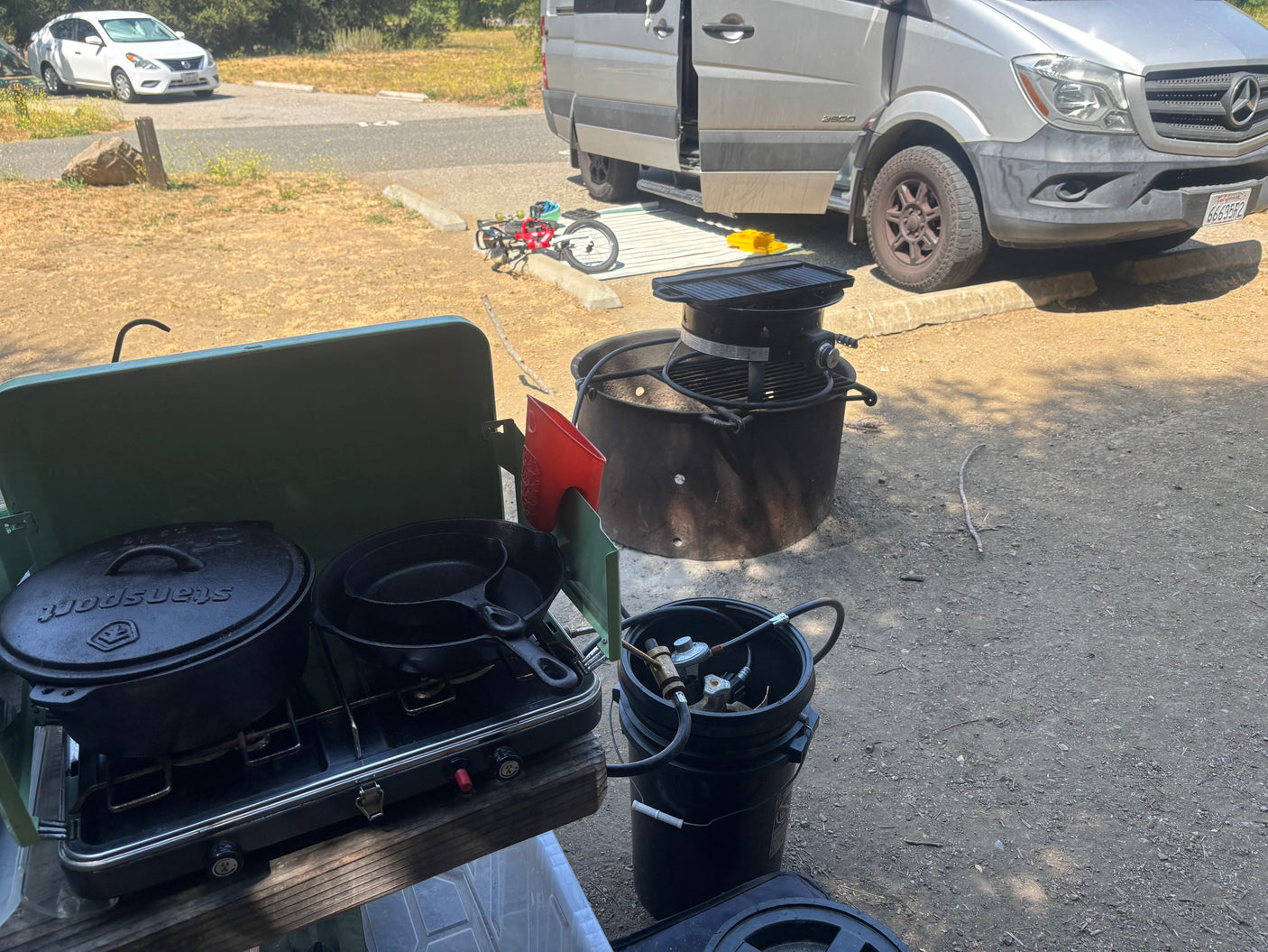 Campsite outdoor cooking setup with RV rental: Cast iron pots on propane stove, fire pit, and Sprinter van nearby.