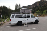 White camper van with a rooftop tent and ladder, parked in a scenic mountain lot. Perfect for an rv rental adventure!