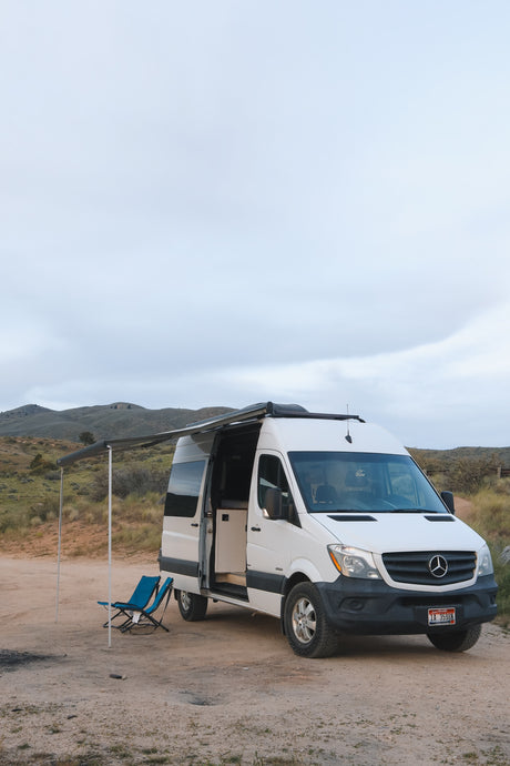 White Mercedes Sprinter camper van with extended awning & chairs on a dirt road by hills. Ideal RV rental for outdoor adventures.