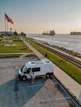 White RV rental van at a scenic waterfront park. A cargo ship sails past while joggers enjoy the sunset view.