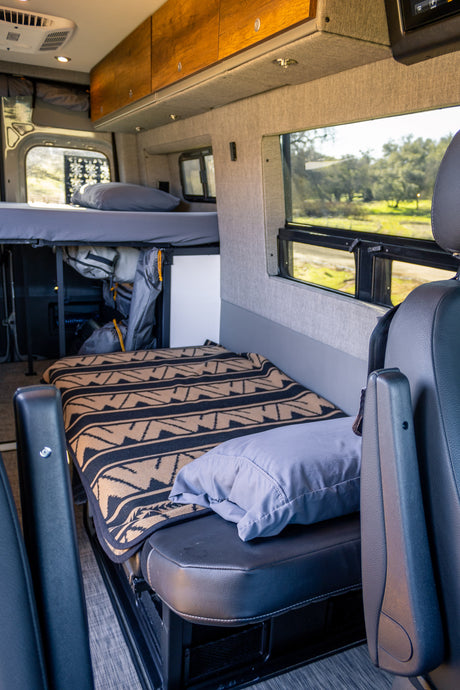 Spacious rv rental interior featuring a bunk bed over storage, a lower bed with a patterned blanket, and scenic window views.