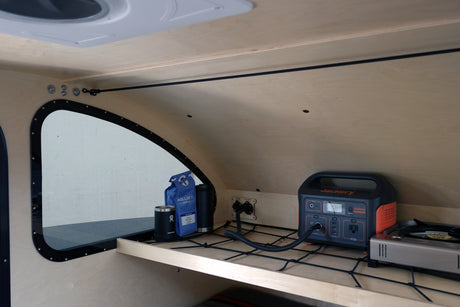 RV rental interior: A wooden shelf stores a Jackery power station, coffee, and a camp stove, secured by a bungee net.