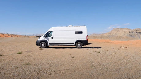 An off-grid RV rental van sits in a wide desert with orange dunes and mountains under a clear sky.