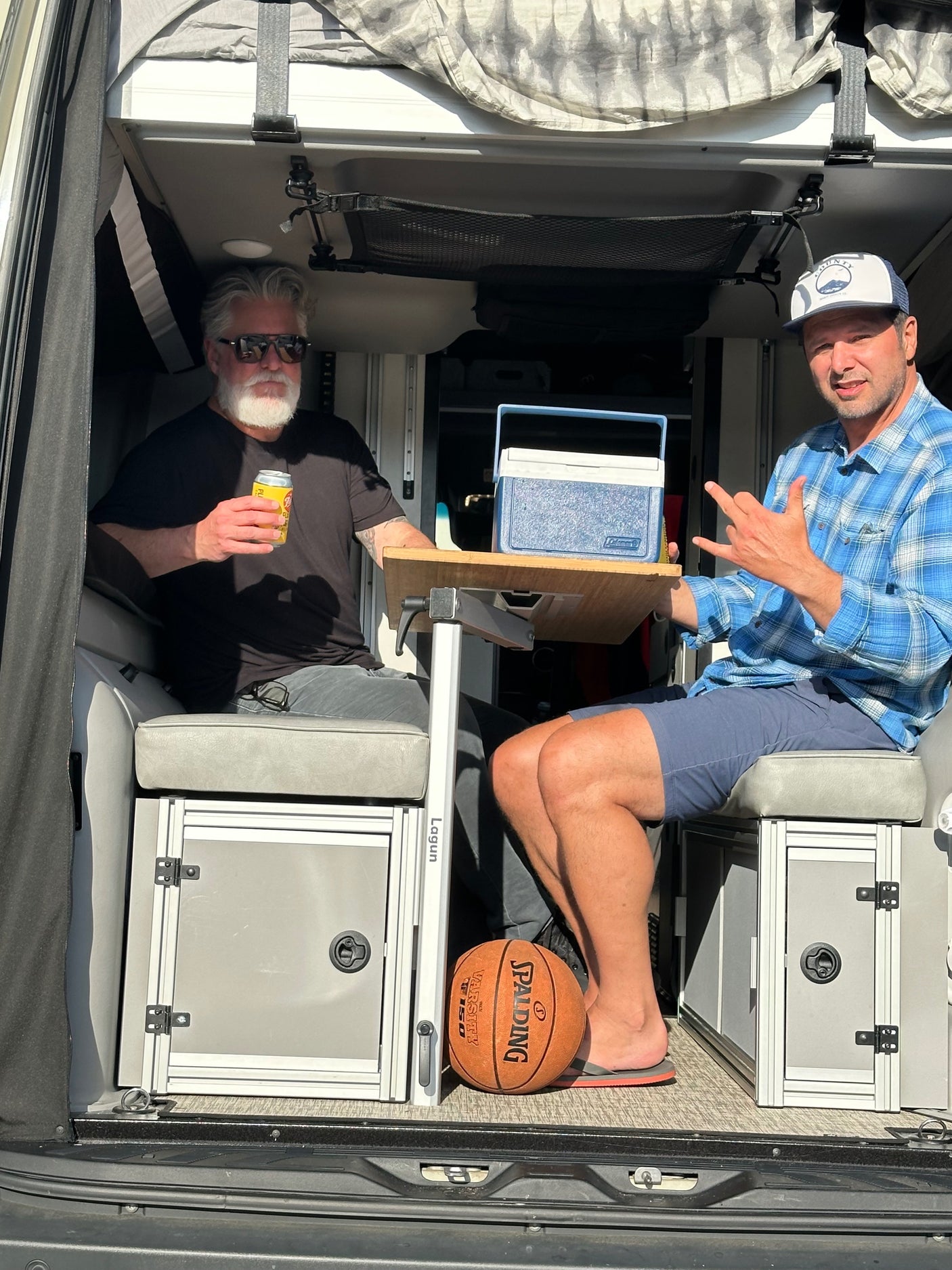 Two friends relax in an RV rental interior with a cooler on the table and a basketball nearby, enjoying their van life adventure.