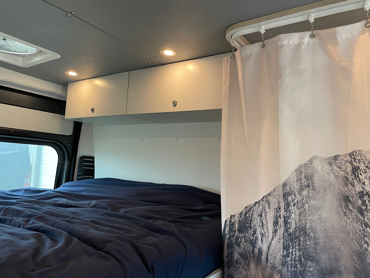 Inside an RV rental: cozy bed with blue bedding, overhead cabinets with lights, and a stylish mountain curtain. — photo 14