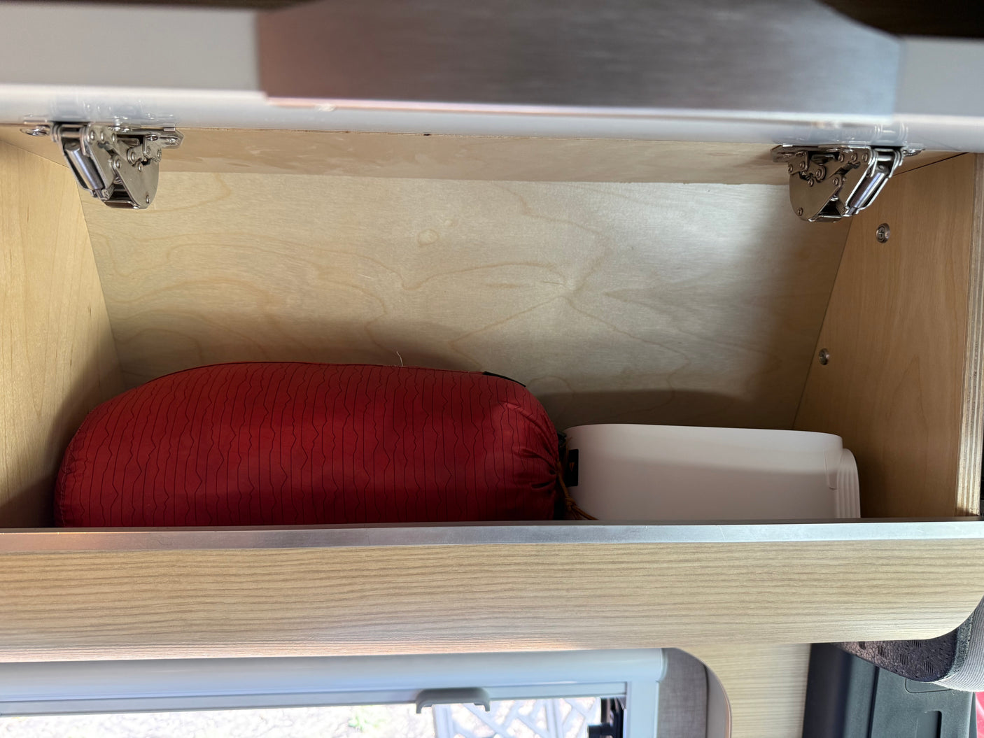 RV rental: An overhead wooden cabinet, seen from inside, holds a red sleeping bag and white container. Visible metal hinges.