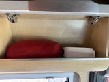 RV rental: An overhead wooden cabinet, seen from inside, holds a red sleeping bag and white container. Visible metal hinges.