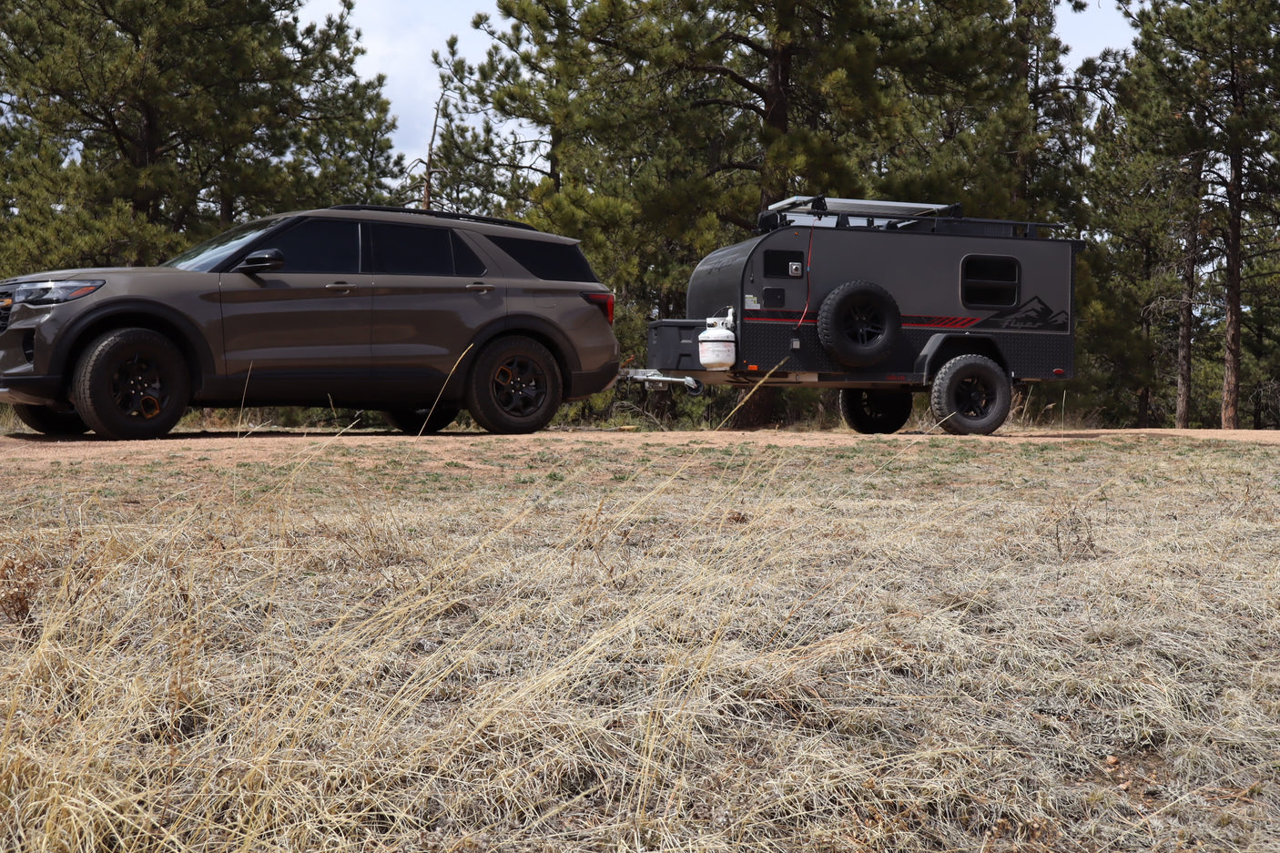 Dark brown SUV towing a grey RV rental camper on a dirt path, surrounded by pine trees and dry grass. Ready for off-road adventures.