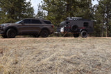 Dark brown SUV towing a grey RV rental camper on a dirt path, surrounded by pine trees and dry grass. Ready for off-road adventures.