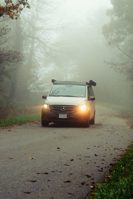 A white RV rental van drives through dense autumn fog on a tree-lined road, its headlights glowing. Serene, misty travel scene.