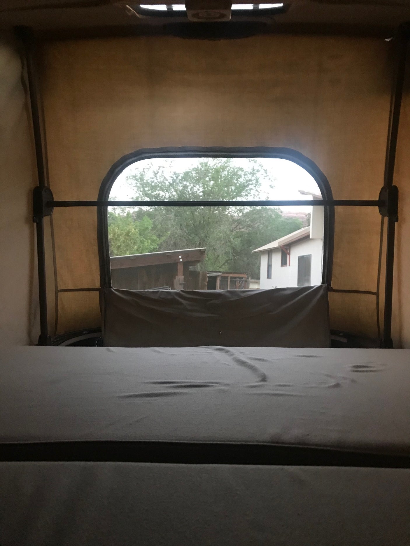 Inside an RV rental rooftop tent, a comfy bed and arched window view of trees and buildings await your camping trip.