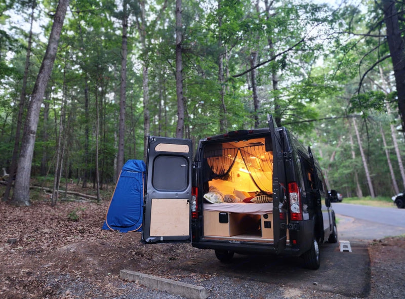 Cozy camper van RV rental set up for camping in a forest. Rear doors open reveal a bed and a blue privacy tent nearby. — photo 12