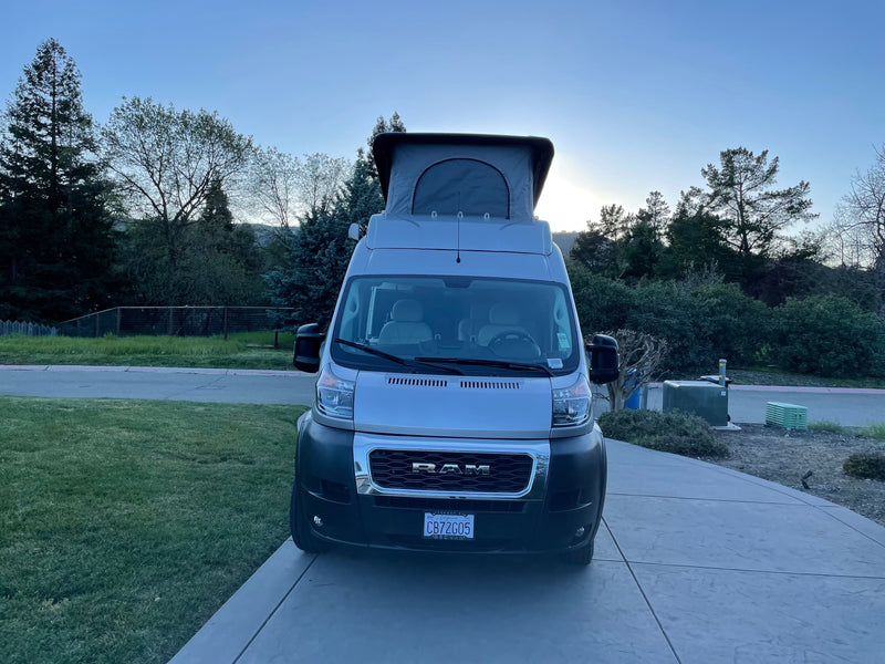 Front view of a light gray Ram ProMaster camper van with a raised pop-top, parked on concrete. Explore with an RV rental! — photo 12