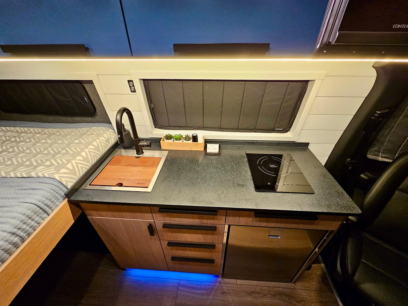 RV rental interior: a compact kitchen with a sink, induction cooktop, and fridge, beside a bed, all under LED lighting. — photo 8
