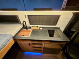 RV rental interior: a compact kitchen with a sink, induction cooktop, and fridge, beside a bed, all under LED lighting.