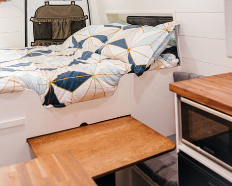 Inside this cozy RV rental, discover a neatly made bed with blue/gold geometric bedding, a wood counter, and clever storage solutions.