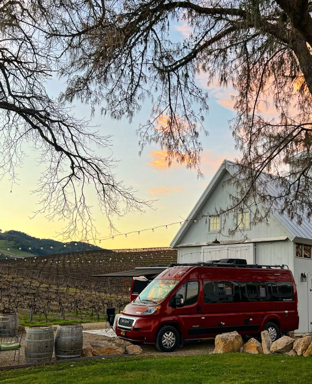  offering scenic desert camping.|A red RV rental parked by a white barn amidst a vineyard at sunset. Bare trees and string lights overhead create a cozy scene.|Two red Winnebago Travato rv rental camper vans are parked side-by-side — photo 2