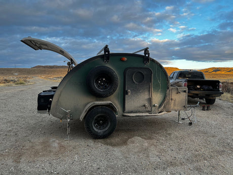 A muddy green teardrop camper with an open rear hatch and truck on a dirt road. Ideal for an rv rental adventure.