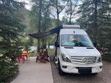 A white rv rental camper van with an extended awning at a scenic riverside campsite with outdoor seating and trees.