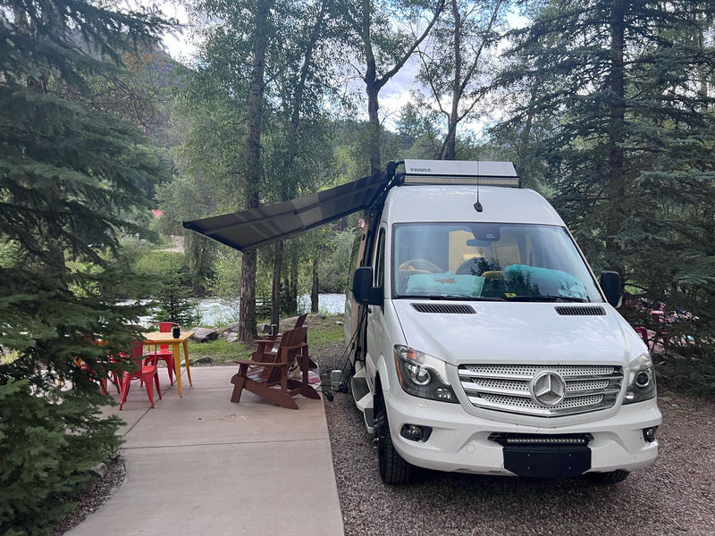  surrounded by forests & distant peaks.|A white rv rental camper van with an extended awning at a scenic riverside campsite with outdoor seating and trees.|White customized Sprinter van RV rental — photo 4