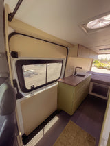 Inside an RV rental: a modern camper van interior with a sleek kitchenette, green cabinets, and a cozy bed. Views from the large window.