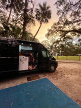 Scenic RV rental camping at sunset. A child is visible inside the black camper van, parked amidst trees.