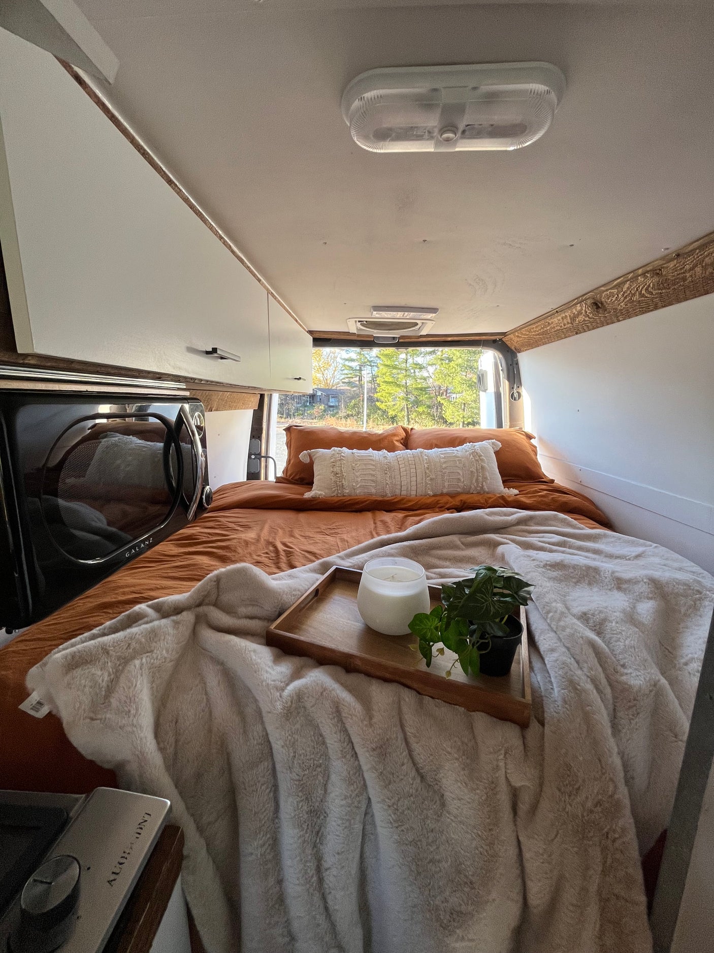 Cozy RV rental bedroom with warm orange bedding, plush white blanket, microwave, and green outdoor view.