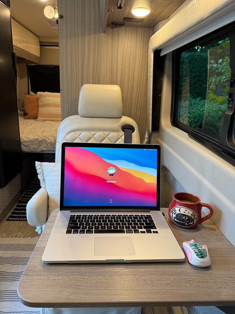 RV rental mobile office: MacBook, mug, & mouse on a table in a cozy camper, with a bed & nature view. Ideal for van life.