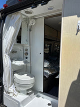 Inside an RV rental: a compact wet bath with toilet, foldable sink, and shower, adjacent to the bed.