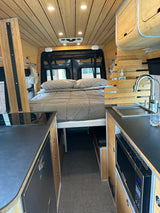 Cozy bamboo-finished RV rental interior featuring a comfortable bed, compact kitchen with sink, stovetop, and microwave.