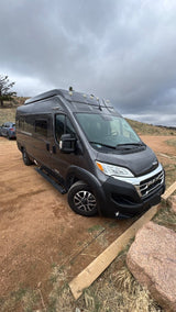 This rugged rv rental camper van, dark grey and ready for exploration, is parked on a dirt path under an overcast sky.