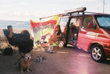 Beach camping with a red rv rental van, awning extended. People relax in chairs, a dog sits on sand by the ocean.