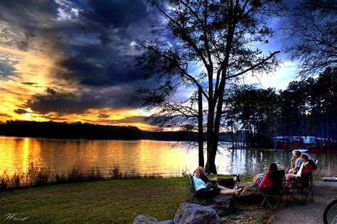 Friends relax at a lake sunset. RV rental makes tranquil lake camping possible. RVs visible across the water.