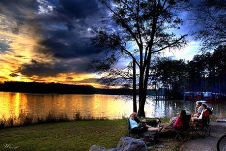 Friends relax at a lake sunset. RV rental makes tranquil lake camping possible. RVs visible across the water.