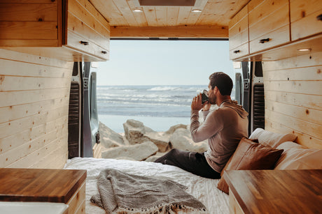 Relaxing in an RV rental! Man sips coffee with stunning ocean views from his cozy, wood-paneled camper van.