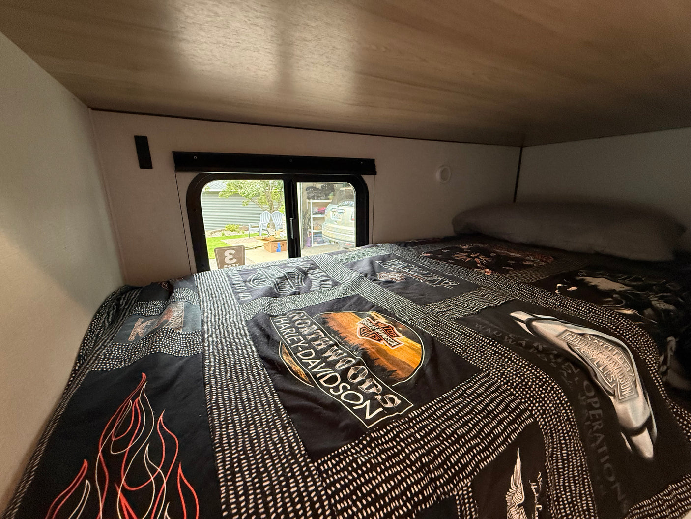 Cozy RV rental bed with a Harley-Davidson quilt, offering a window view of a residential area.