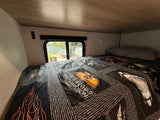 Cozy RV rental bed with a Harley-Davidson quilt, offering a window view of a residential area.