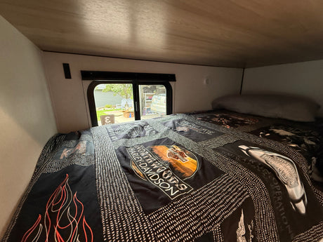 Cozy RV rental bed with a Harley-Davidson quilt, offering a window view of a residential area.