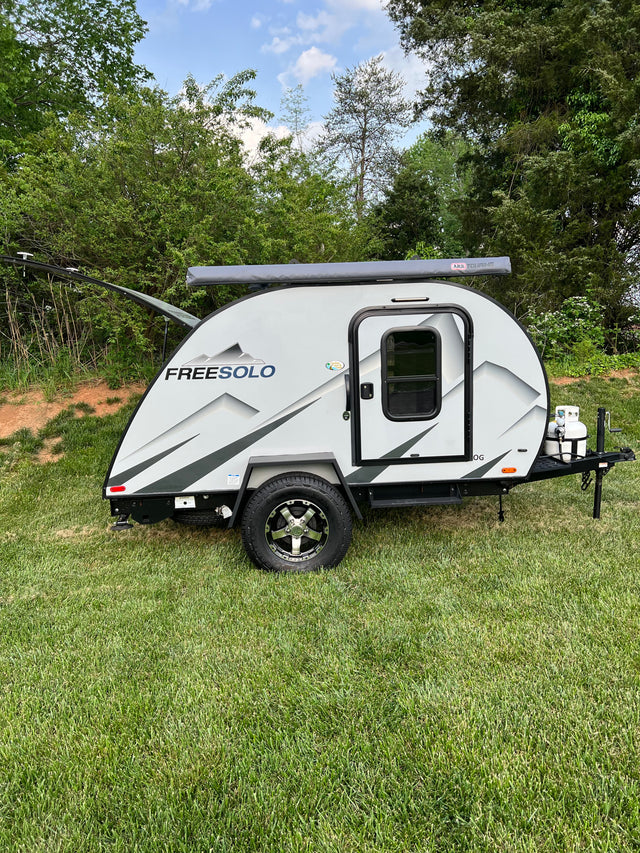This Freesolo teardrop camper RV rental is grey with black accents, an awning, and propane tank. Parked on green grass.