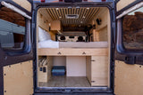 Inside a custom-built camper van, showing a bed, wood paneling, and storage options. Ideal for an rv rental.