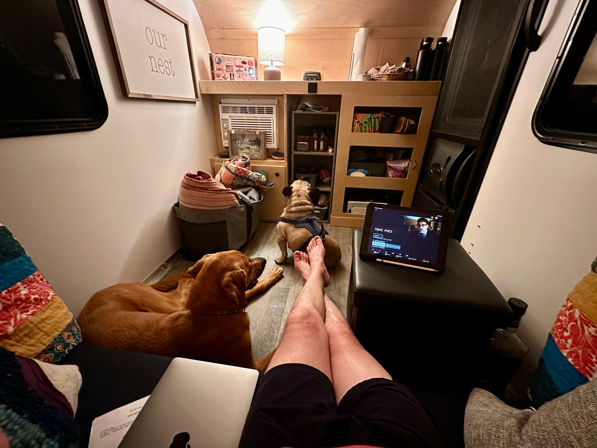 Cozy RV rental interior. Person's legs up, laptop & tablet (Three Pines playing), two dogs relaxing. "Our nest" sign above. — photo 12